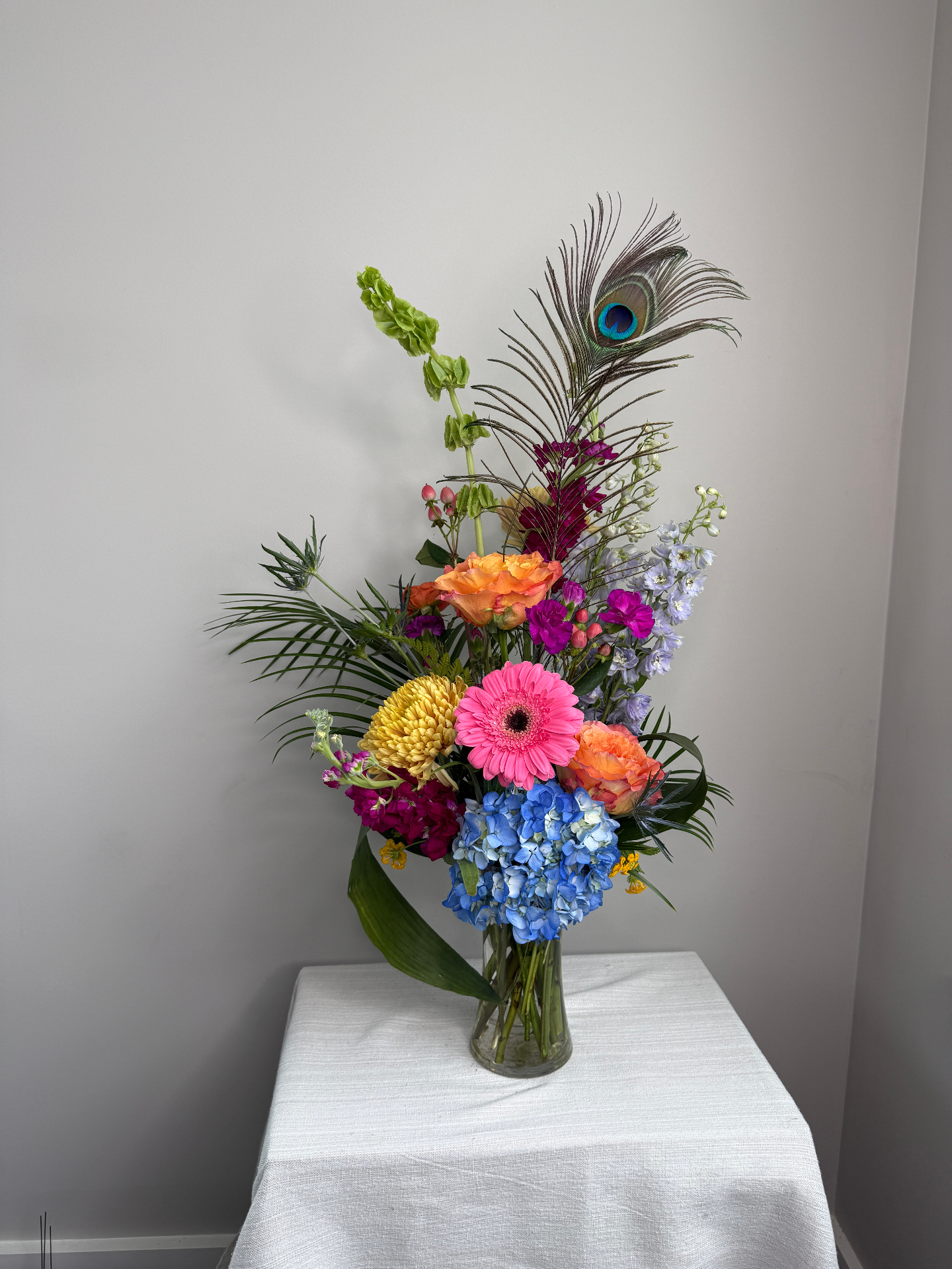 Tall celebration arrangement with a peacock feather, gerbera daisy, and blue hydrangea by Dolce Flowers in Greenville, SC