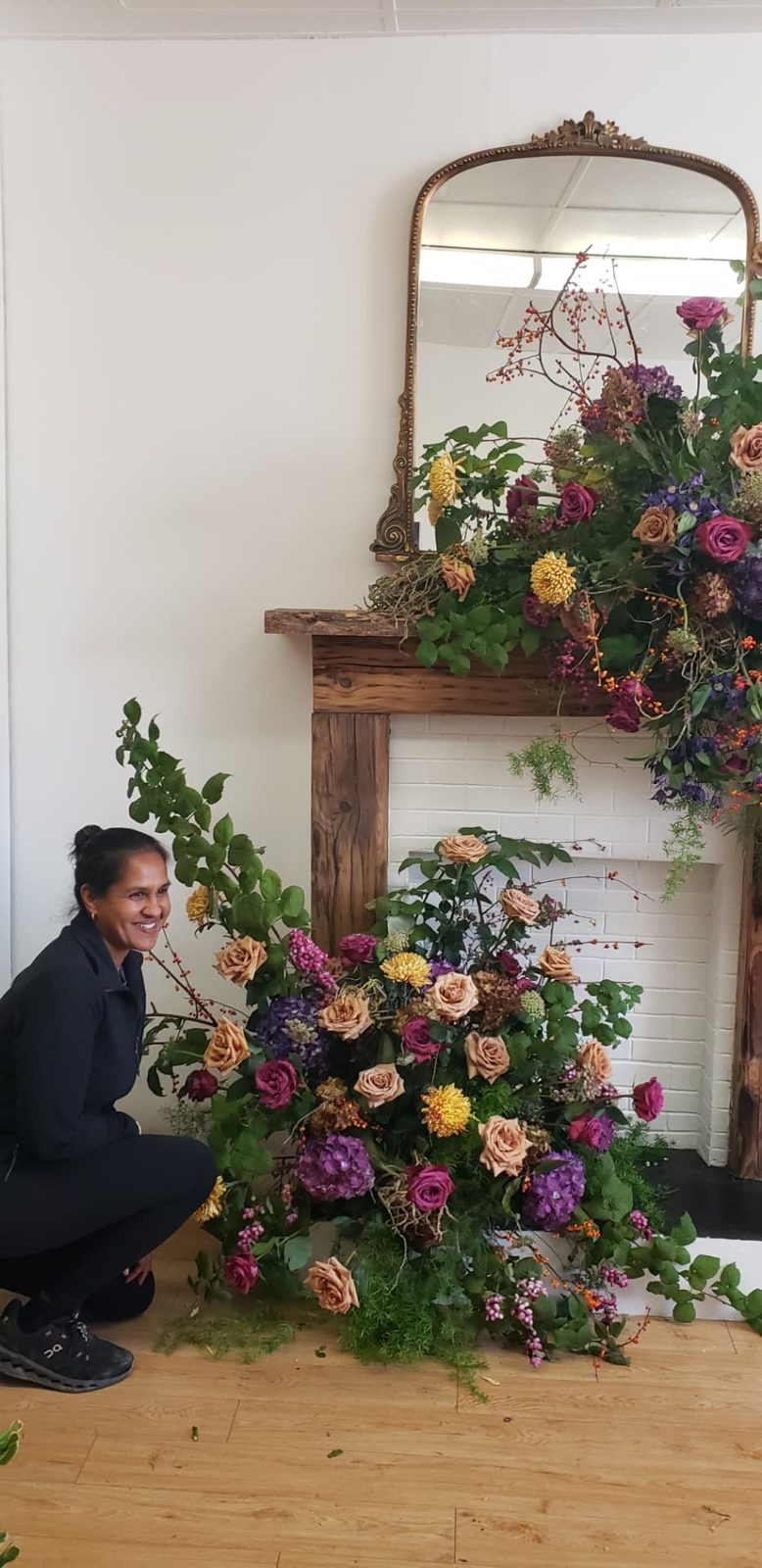 Kara of Dolce Flowers kneeling beside an autumn mantel installation of toffee roses, purple hydrangea, and bittersweet vine